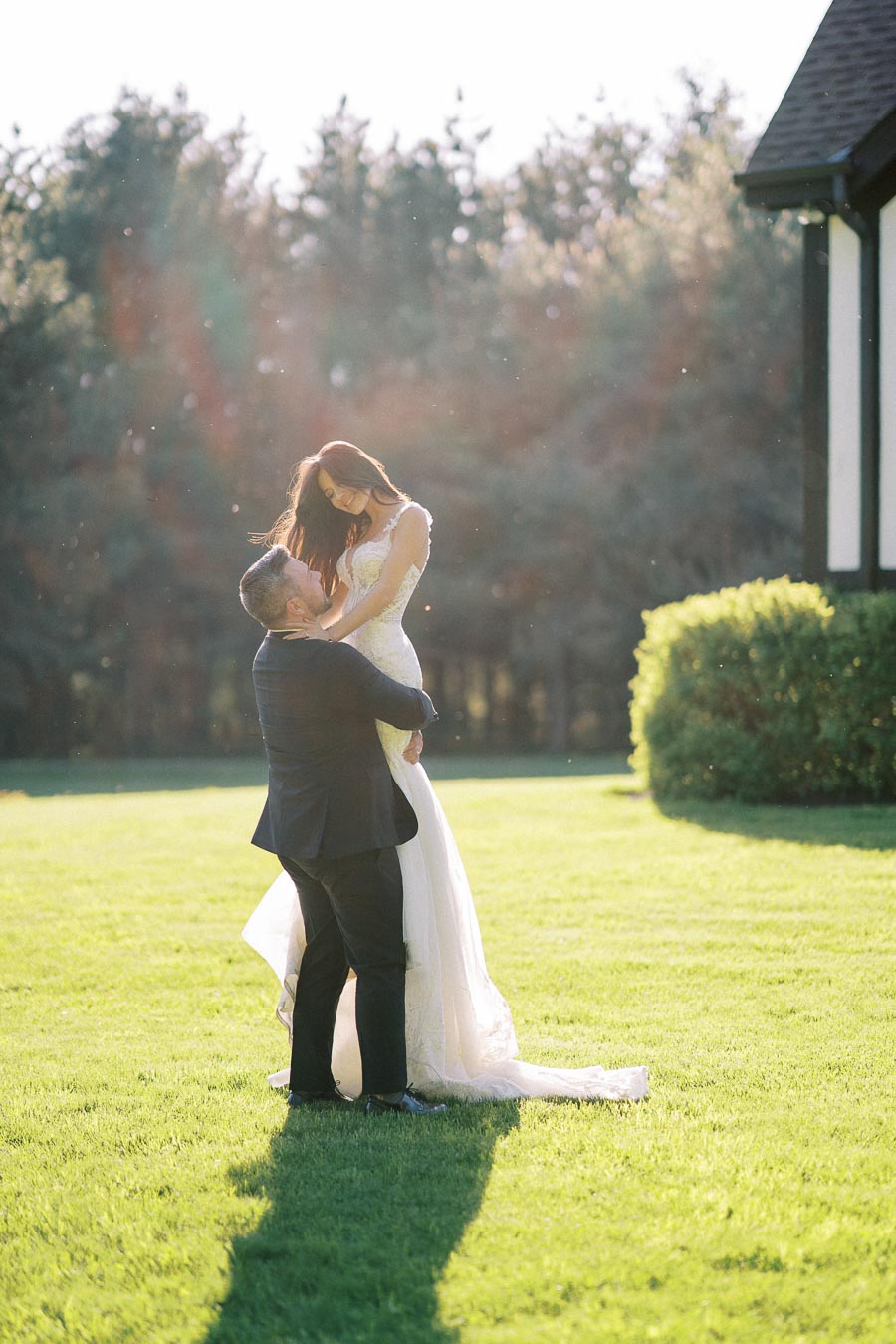 A bride and groom share a joyful moment outdoors on a sunny day, with the groom lifting the bride in a beautiful embrace on a green lawn, surrounded by lush trees and elegant architecture.