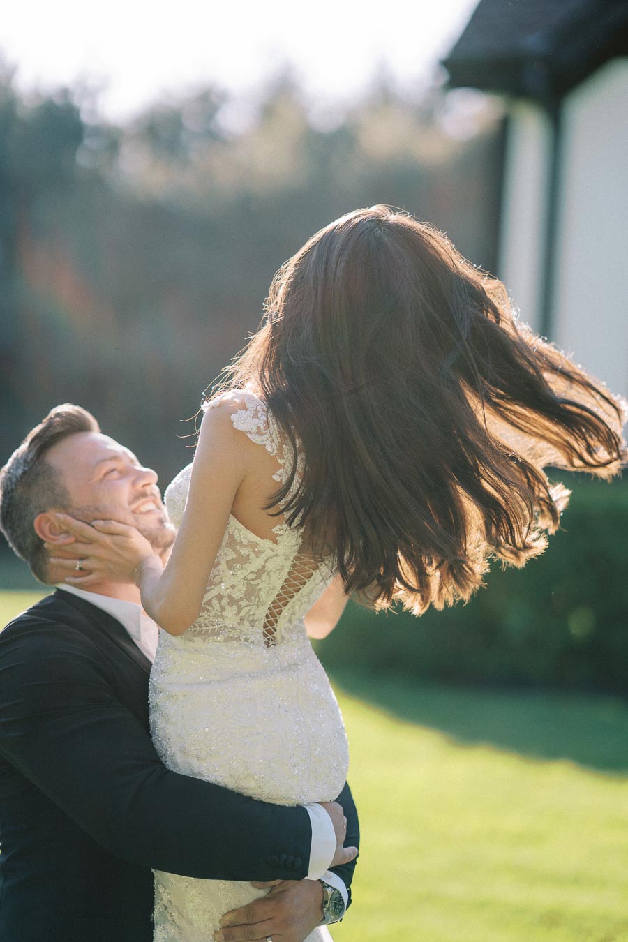 A happy couple embracing outdoors on a sunny day, with the woman in a beautiful lace wedding dress being lifted by her partner wearing a suit, surrounded by lush greenery.