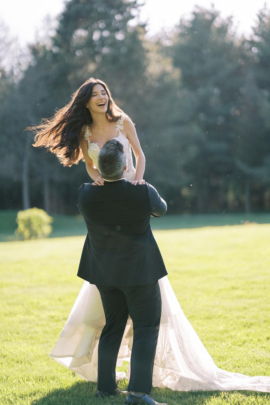 A groom playfully lifts a laughing bride in a joyful outdoor wedding moment, set against a backdrop of lush greenery and soft sunlight.