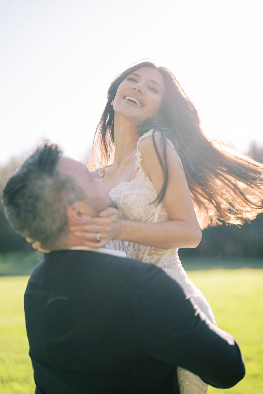 A joyful bride in a lace wedding dress being lifted by her partner outdoors, both smiling brightly in the sunlight on a green lawn.