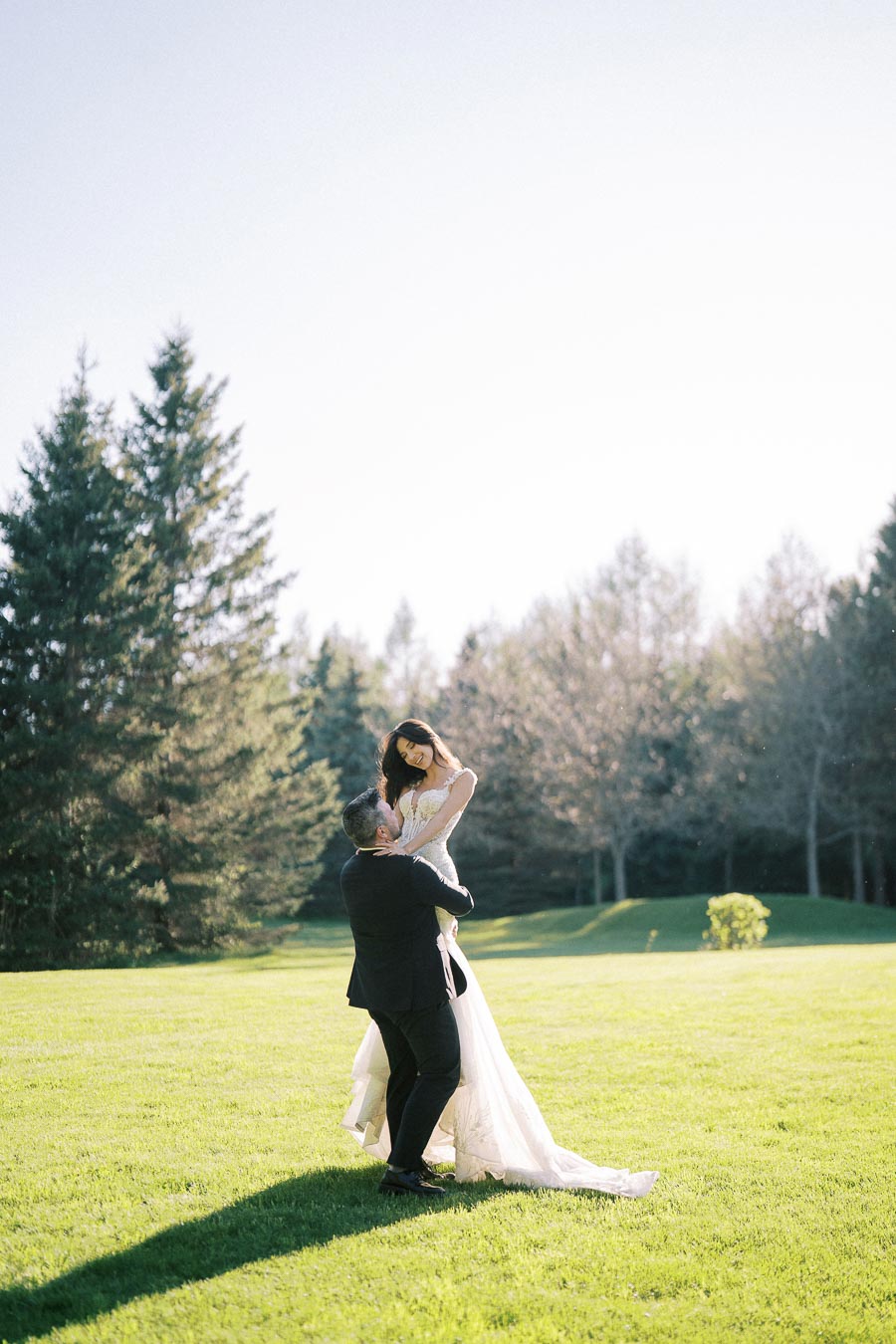 A joyful bride in a white wedding dress is lifted by the groom in a black suit on a sunlit green field with tall evergreens in the background.