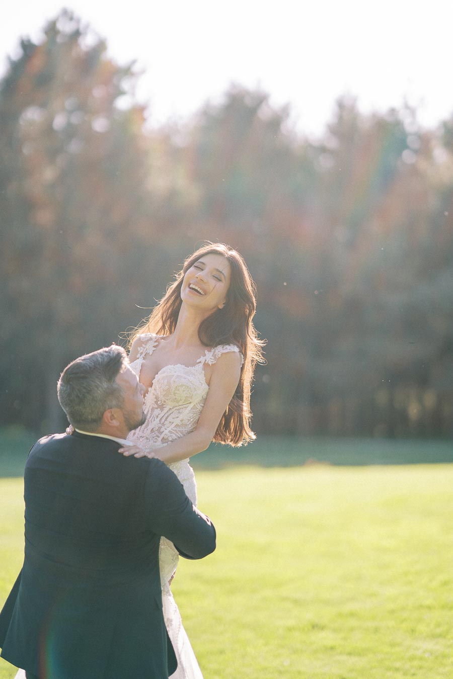 A joyful couple celebrating their wedding day in a sunny outdoor setting, with the bride laughing as the groom lifts her against a backdrop of trees and a bright, green lawn.