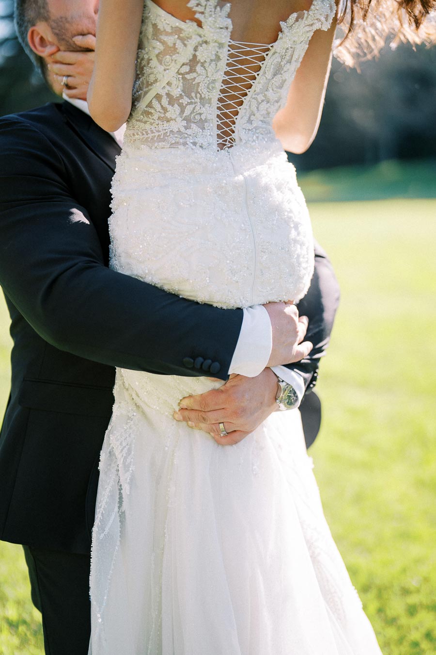 A groom lifts his bride, showcasing the intricate lace and beaded detailing of her wedding dress, in a sunlit outdoor setting.