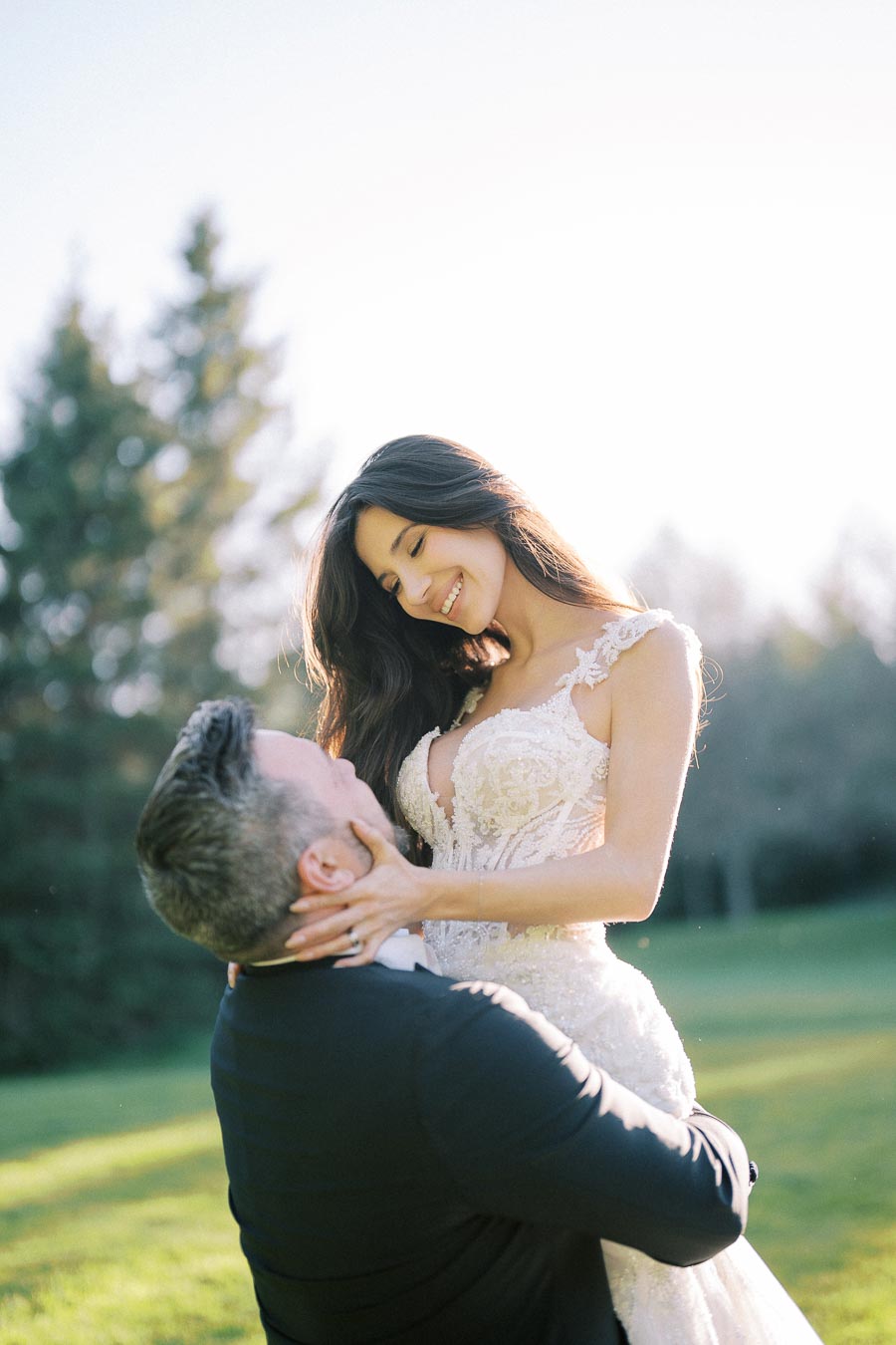 Bride smiling, wearing an elegant lace wedding dress, playfully embraced by groom in a suit, set in a sunlit outdoor scene with trees.