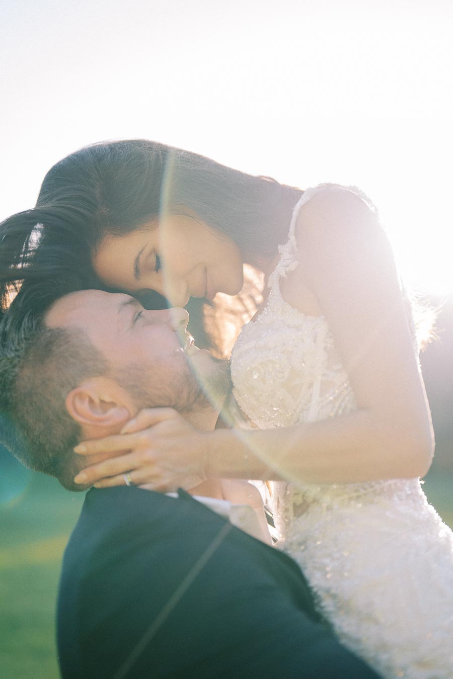 A romantic couple embracing in sunlight, bride wearing a beaded lace gown, outdoor wedding photography.