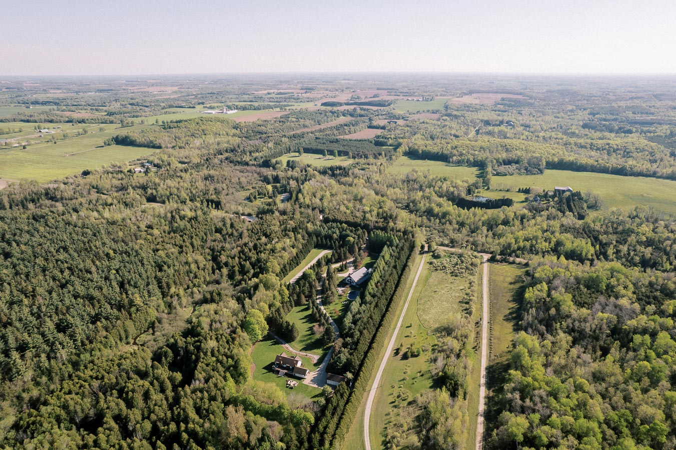Aerial view of scenic rural landscape featuring dense forests, open green fields, and a few residential houses, under a clear blue sky.