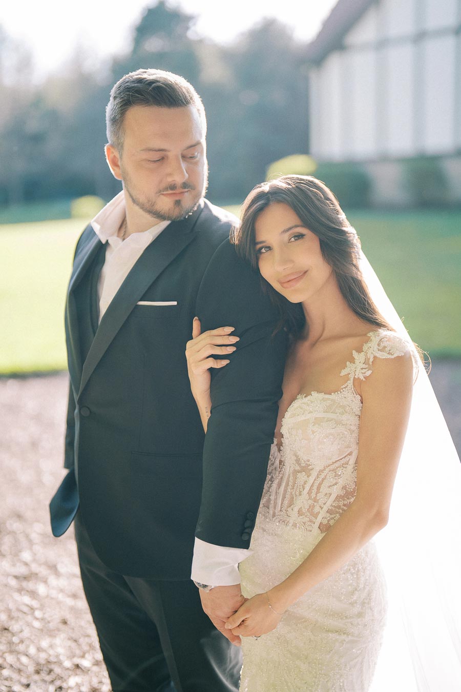 A bride in a lace wedding gown and a groom in a black suit pose outdoors on their wedding day, with a soft sunlight backdrop highlighting their romantic moment.