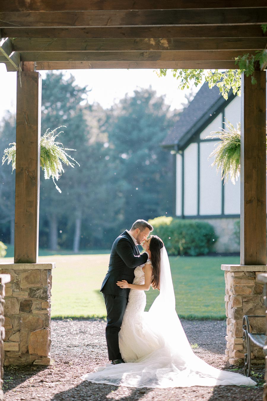 Romantic bride and groom embracing under rustic wooden pergola with greenery, outdoor wedding setting in a sunny garden.