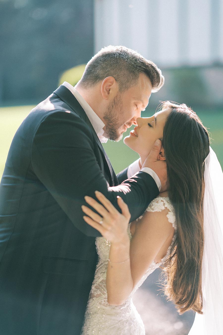 Romantic wedding couple sharing an intimate moment, groom in black suit and bride in elegant white gown with veil, embracing outdoors.