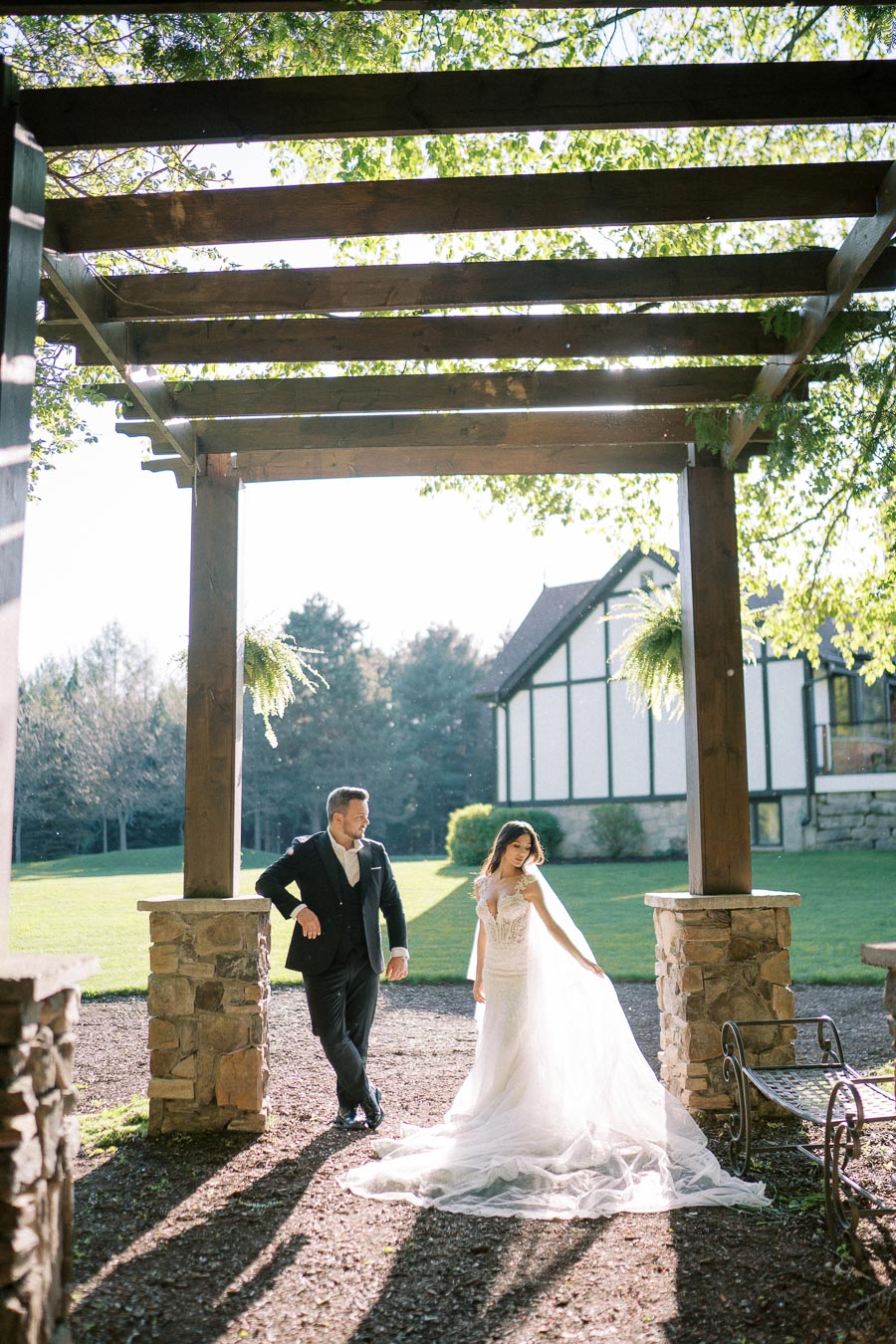 A couple in elegant wedding attire standing under a wooden pergola in a garden setting with a Tudor-style building in the background, surrounded by lush greenery and dappled sunlight.