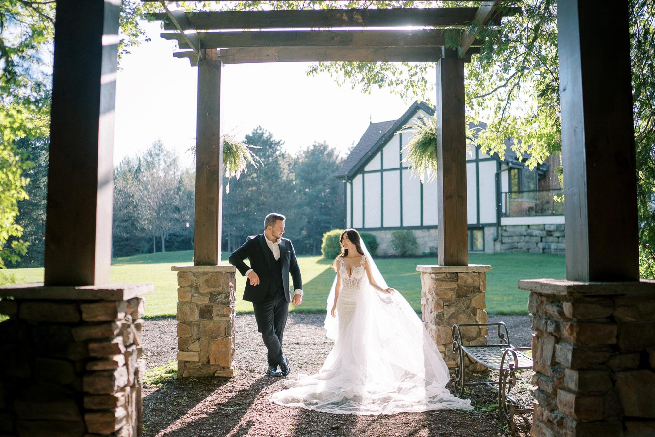Elegant bride and groom at outdoor wedding venue, framed by rustic stone archway and lush garden backdrop