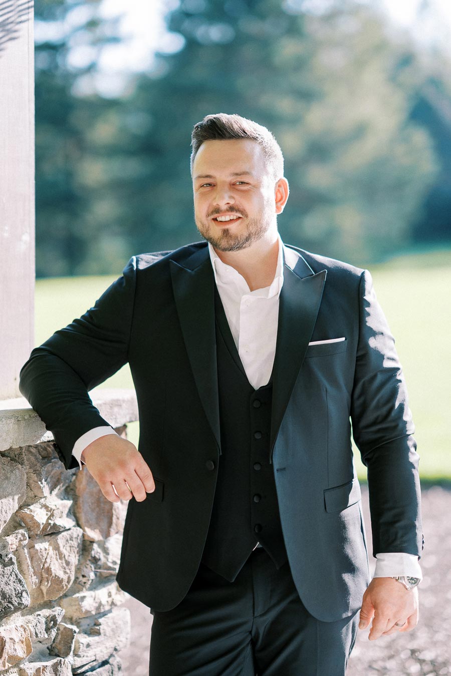 Man in a formal black suit standing by a stone wall with a blurred green outdoor background, smiling confidently.