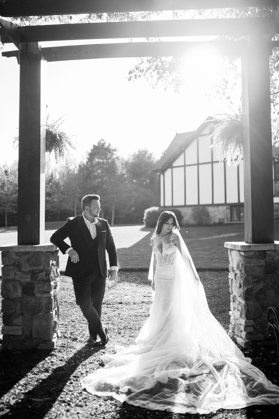 Black and white image of a bride in an elegant wedding gown and veil walking alongside a groom in a suit under a rustic wooden pergola, with a scenic backdrop of a Tudor-style house and sunlit garden.