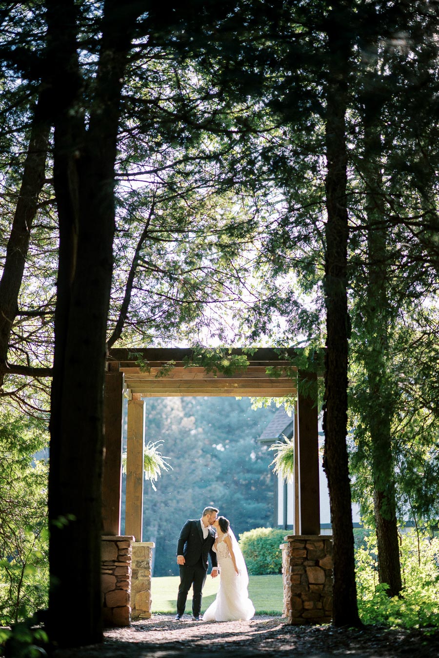 A bride and groom share a kiss under a wooden archway surrounded by lush greenery and tall trees, creating a picturesque forest wedding setting.