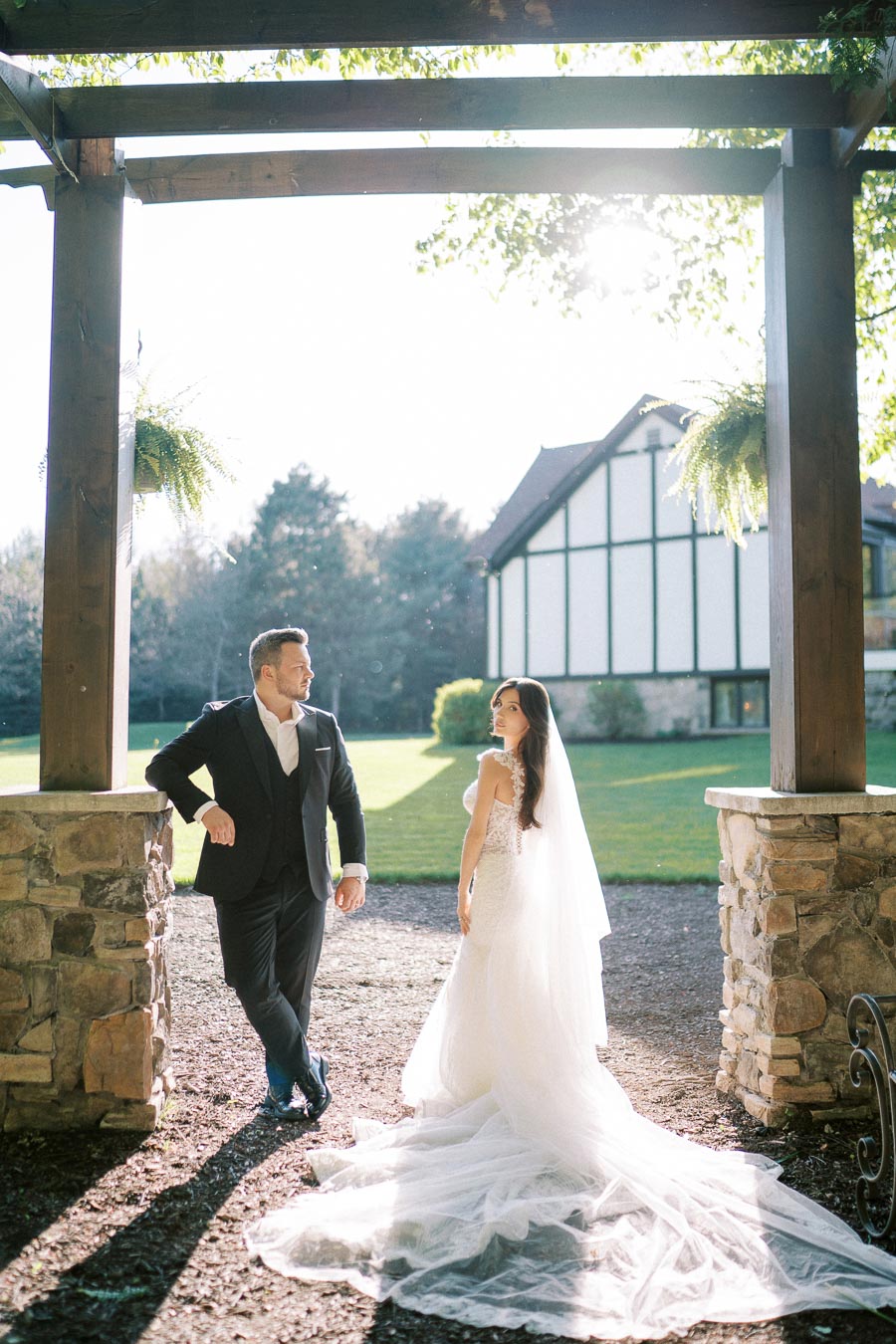 Elegant bride and groom standing under a wooden pergola with sunlight filtering through, next to a stone house with a lush green lawn.