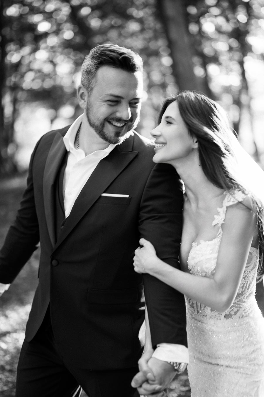 Black and white photo of a joyful couple walking hand in hand through a sunlit forest, with the woman wearing a lace wedding dress and the man in a suit, capturing a moment of love and happiness.