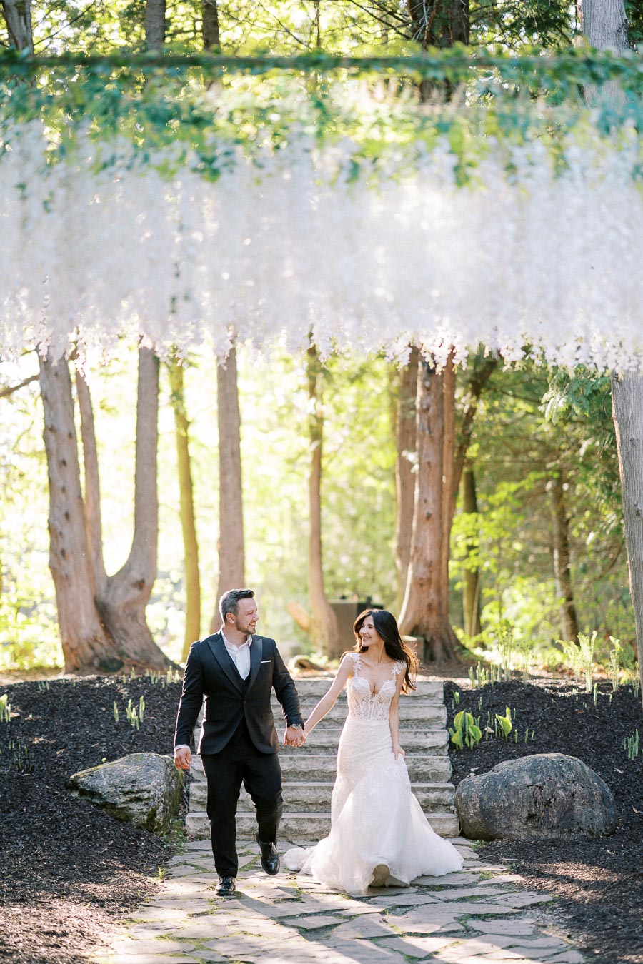A newlywed couple joyfully holding hands and walking down stone steps under a canopy of hanging white flowers, surrounded by lush green trees.