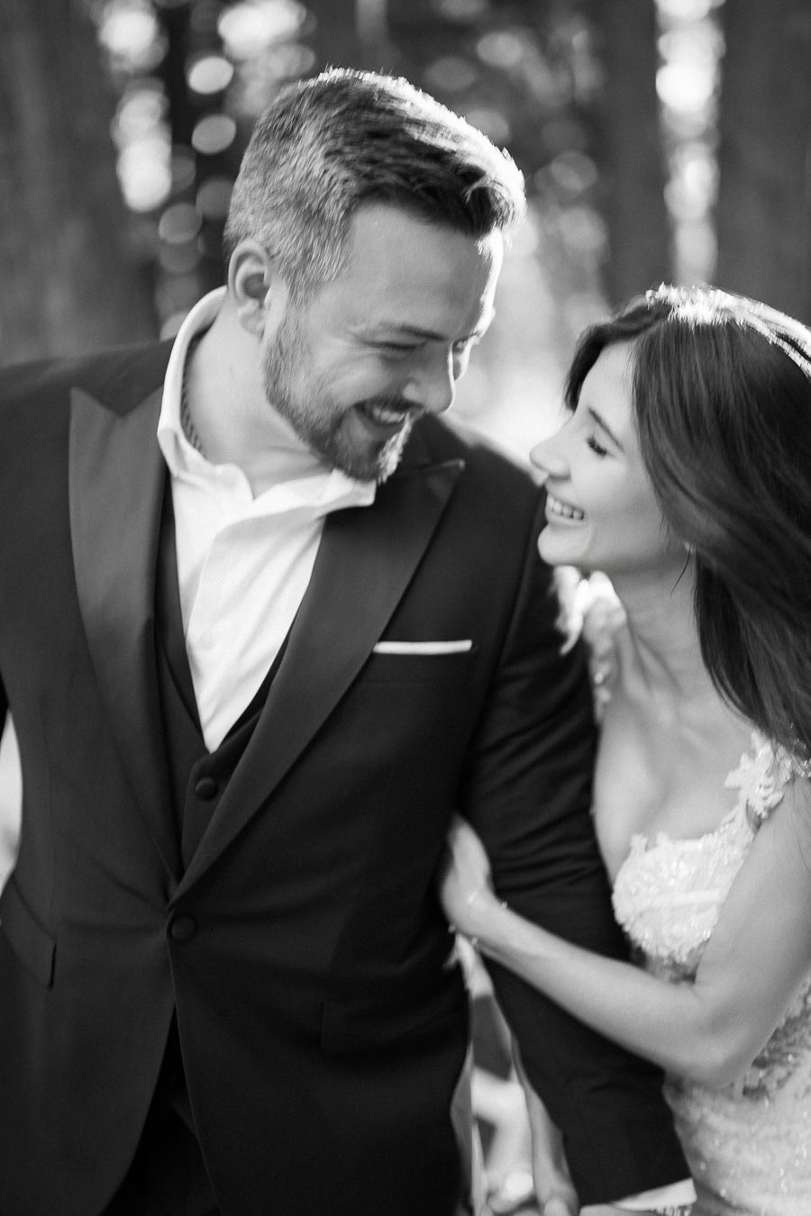 Black and white photo of a joyful couple on their wedding day, with the bride in a lace gown and groom in a suit, sharing a loving smile outdoors.