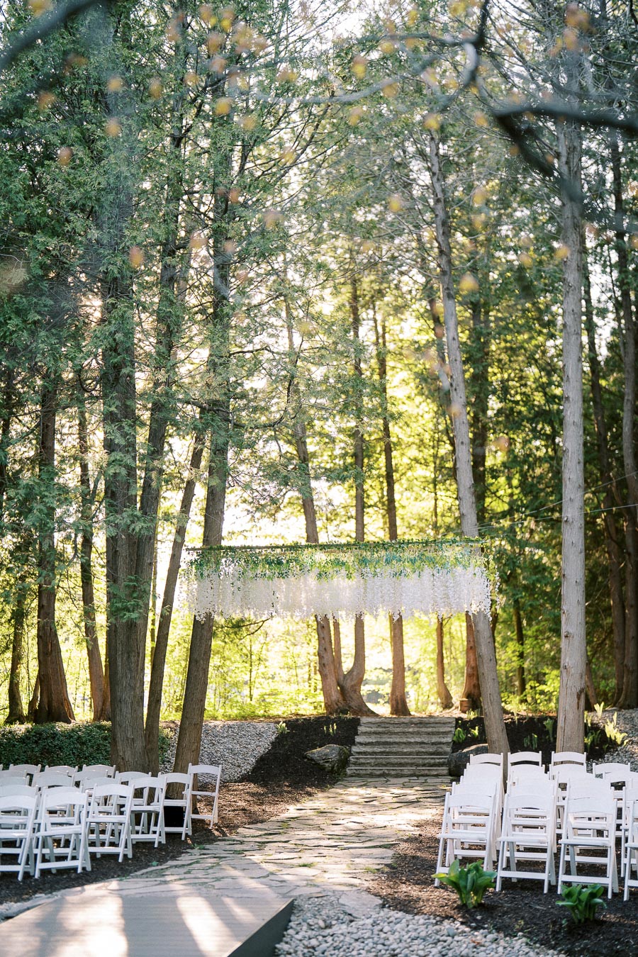 Outdoor wedding ceremony setup in a serene forest setting, featuring white folding chairs arranged on either side of a stone path leading to a floral arch adorned with greenery and white flowers. Sunlight filters through tall trees, creating a peaceful atmosphere.