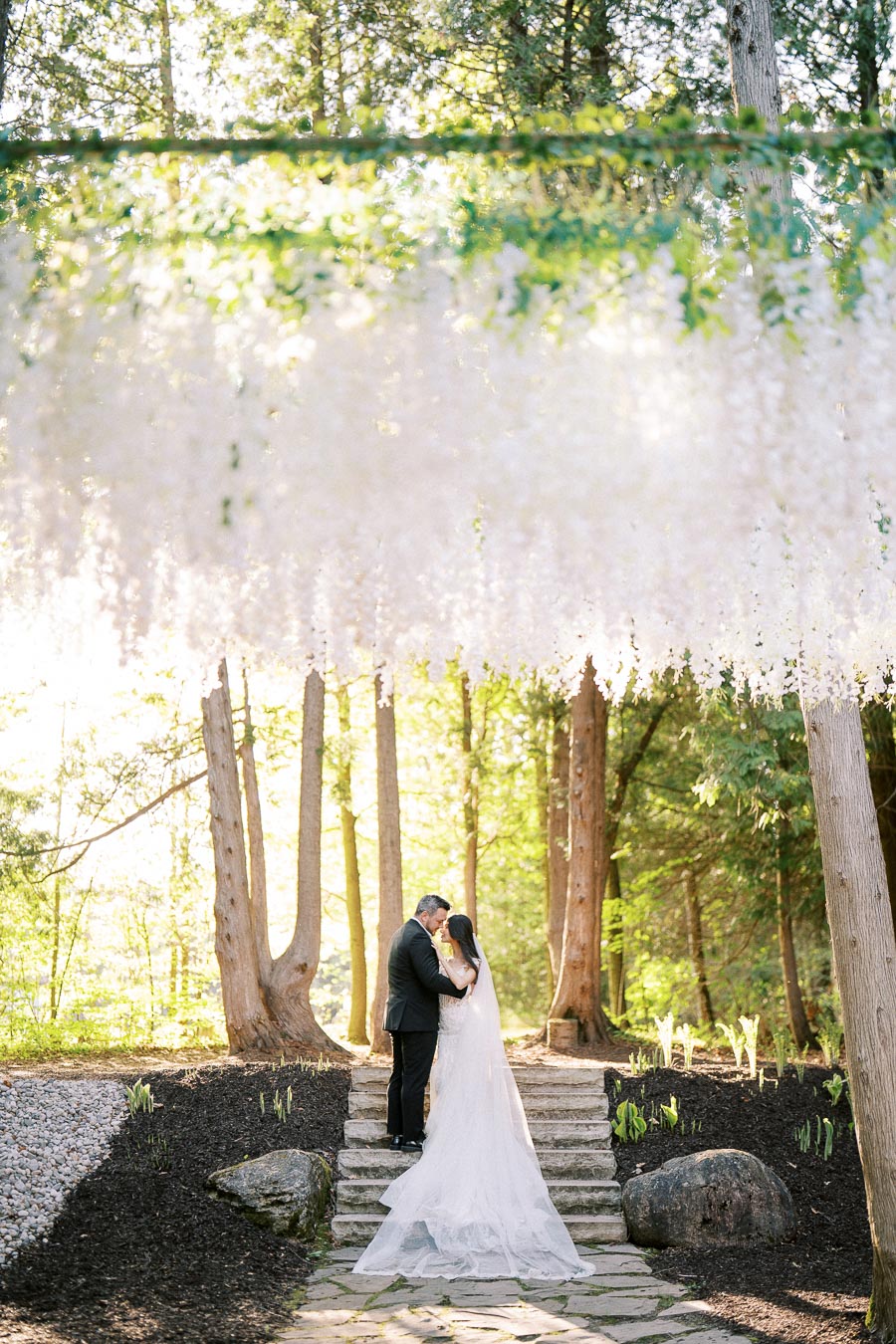 A bride and groom embrace under a canopy of white hanging flowers in a sunlit forest setting, surrounded by tall trees and natural stone steps, creating a romantic and serene atmosphere.