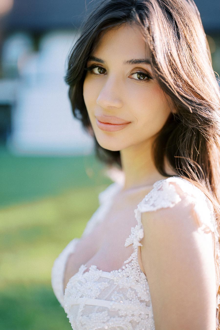 Close-up of a woman with long, dark hair wearing a delicate white lace dress, standing outdoors with a soft, blurred green background.