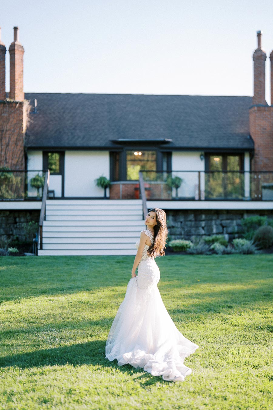 A bride in a flowing white wedding gown stands on a lush green lawn in front of a rustic house with a staircase and brick chimneys, captured on a sunny day.
