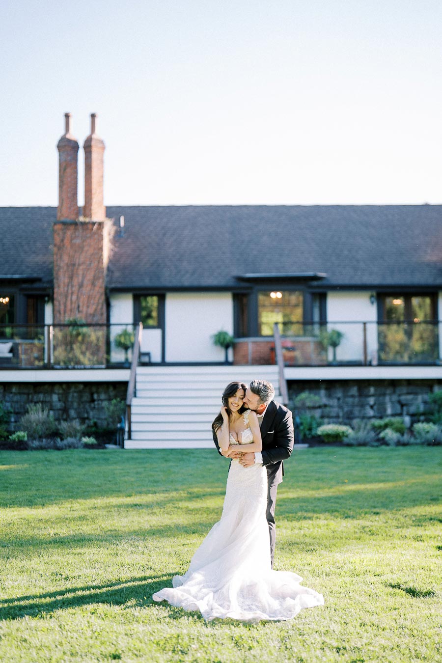 Elegant bride and groom embracing on a green lawn in front of a rustic wedding venue, showcasing love and romance in a serene outdoor setting.