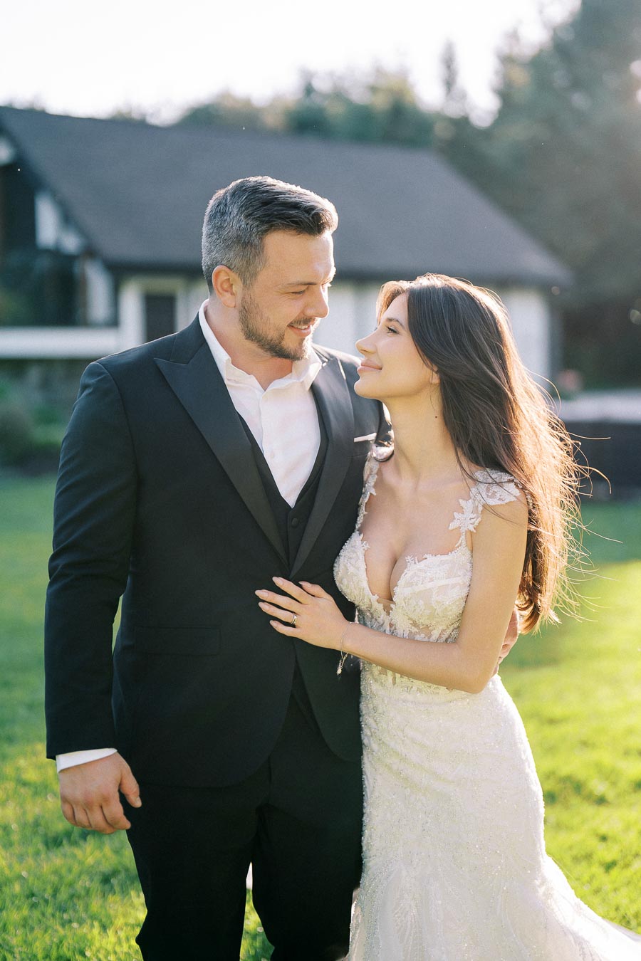 Bride and groom embracing outdoors in sunlight, with bride in a lace wedding dress and groom in a dark suit, smiling at each other, standing in front of a house and lush green lawn.