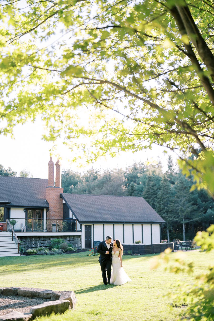 Couple walking on lawn in front of a picturesque house, surrounded by lush greenery and sunlight filtering through the trees, capturing a serene outdoor moment.