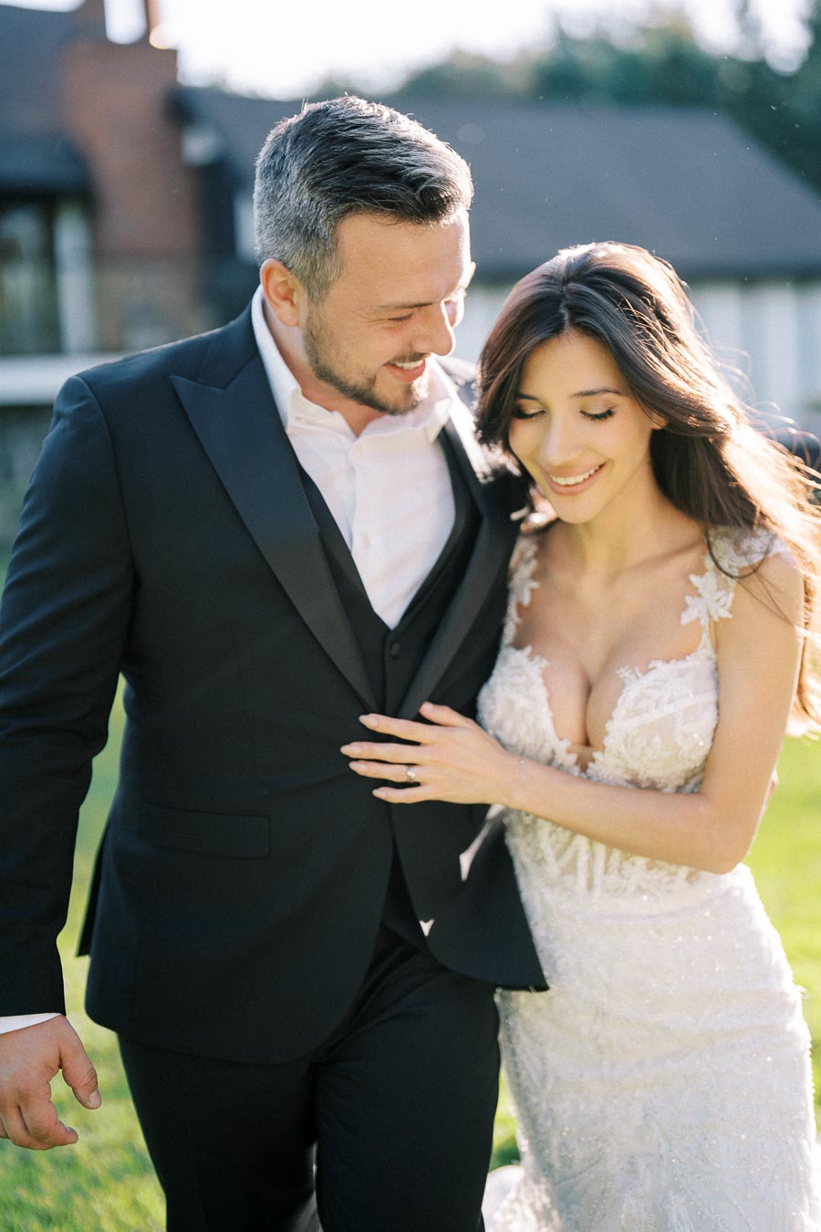 Happy newlywed couple walking outdoors, groom in a black suit and bride in an elegant white lace wedding dress, smiling and sharing a moment of joy on their wedding day.
