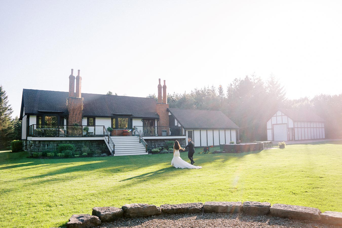 Bride and groom dancing on the lawn of a picturesque estate under a clear sky, surrounded by lush greenery and a large house in the background.