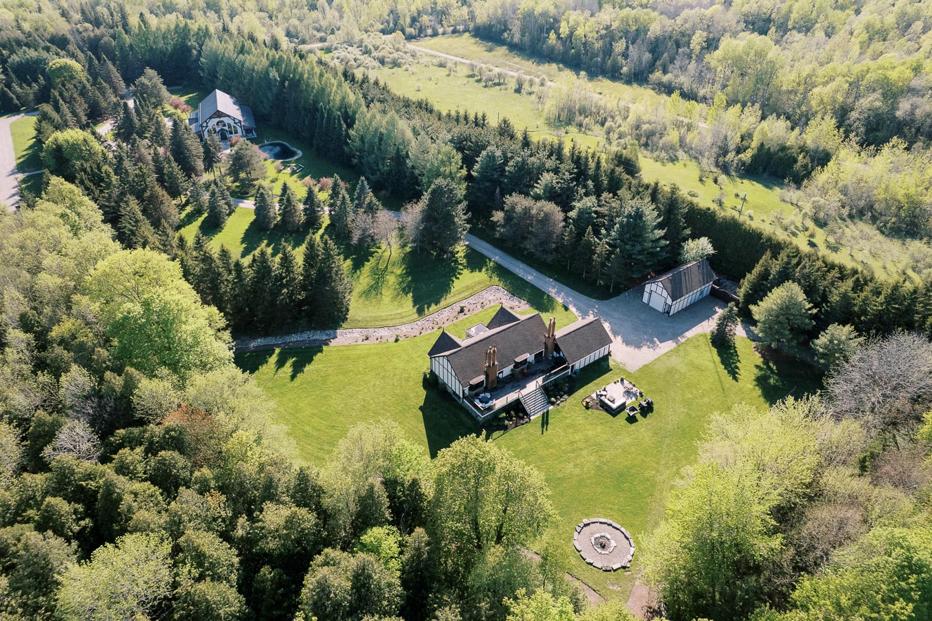 Aerial view of a luxury countryside estate surrounded by lush greenery and dense forest, featuring a large main house, additional buildings, and well-manicured lawns, ideal for a serene and private lifestyle retreat.