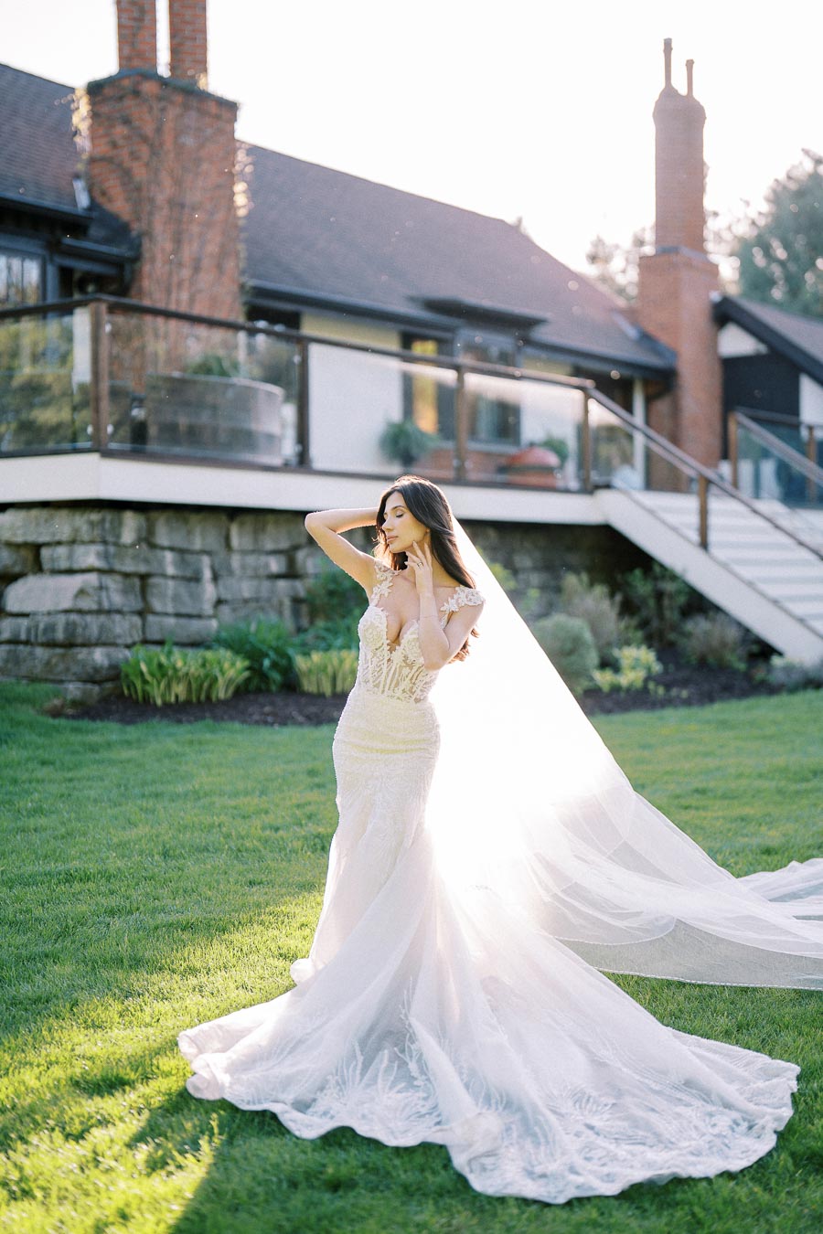 A bride in a flowing white gown poses elegantly on a grassy lawn with a modern house in the background, bathed in warm sunlight.