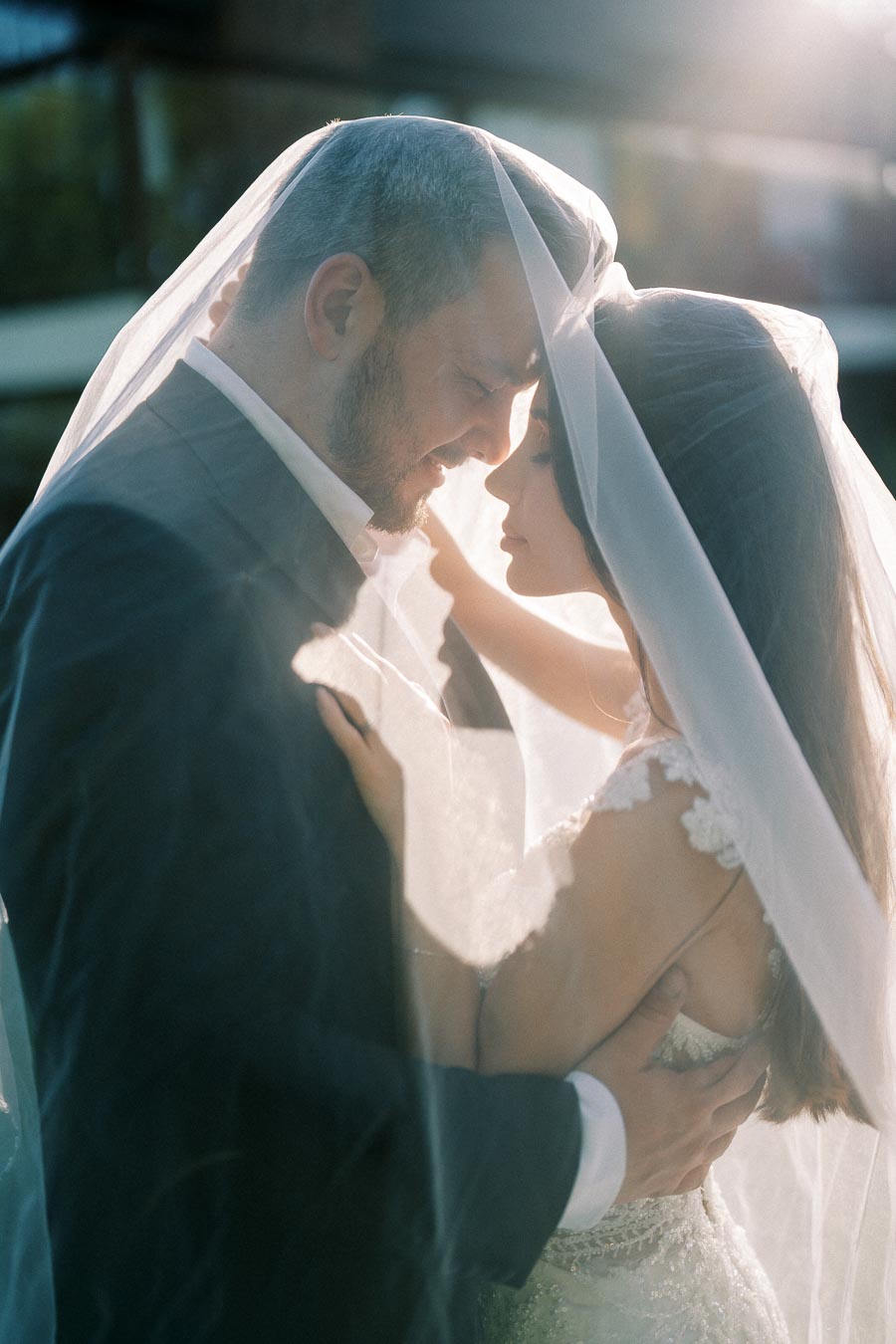 A romantic wedding moment with a bride and groom embracing under a veil, captured in soft sunlight.