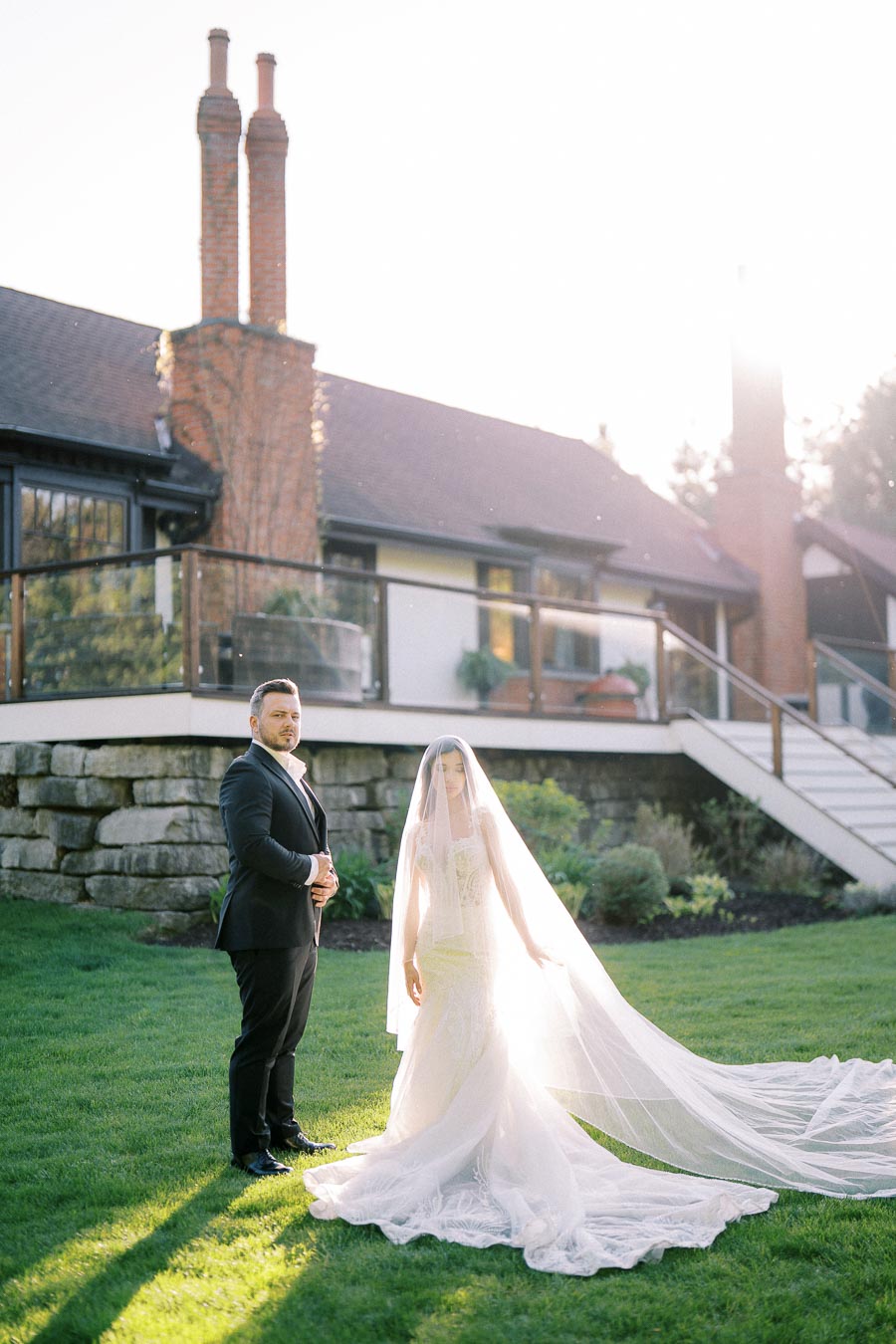 Elegant bride and groom standing on lush green lawn in front of a charming house, with soft sunlight highlighting the bride's flowing veil and lace wedding gown, creating a romantic outdoor wedding scene.