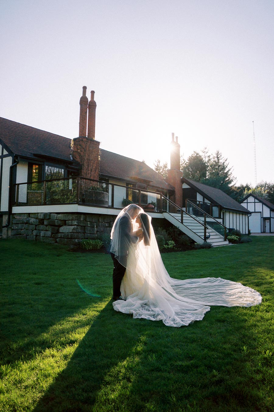 A bride and groom embrace under a flowing veil on a sunlit lawn in front of a charming cottage-style venue, capturing a romantic wedding moment.