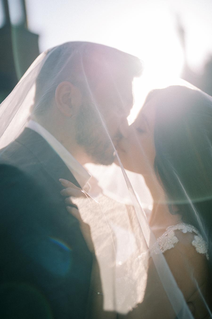 Romantic wedding moment as bride and groom kiss under veil with sunlight flare