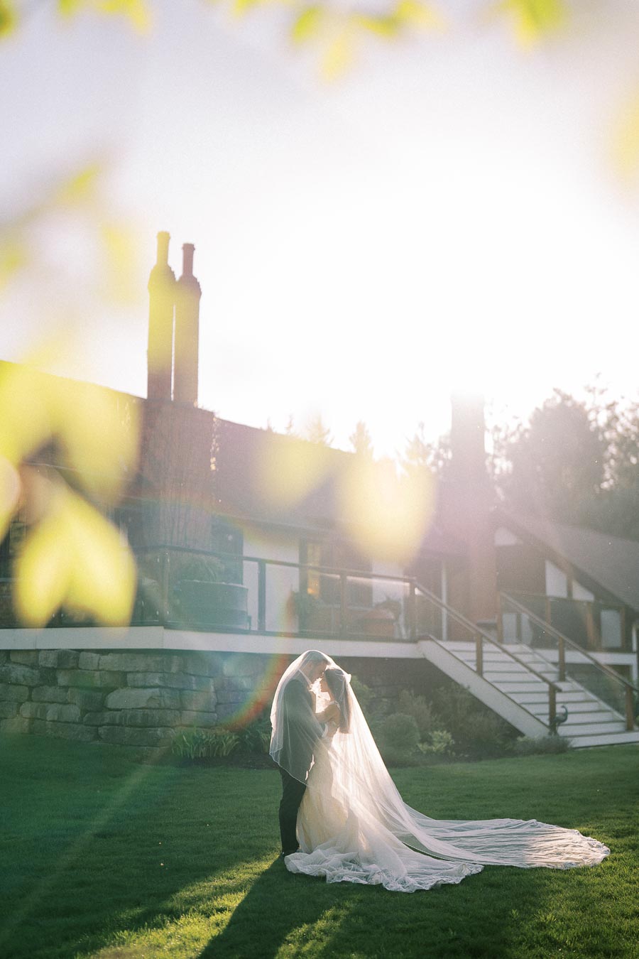 A bride and groom embrace under a long wedding veil on a sunlit lawn in front of a charming house, with sunlight creating a soft, romantic atmosphere.