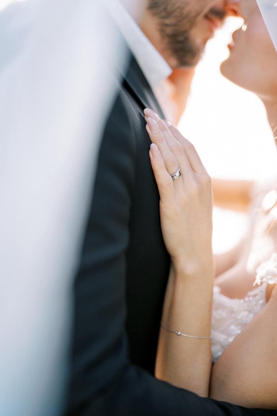 A close-up image of a bride and groom about to kiss, highlighting the bride's hand with a wedding ring, gently resting on the groom's shoulder, set against a soft, bright background.