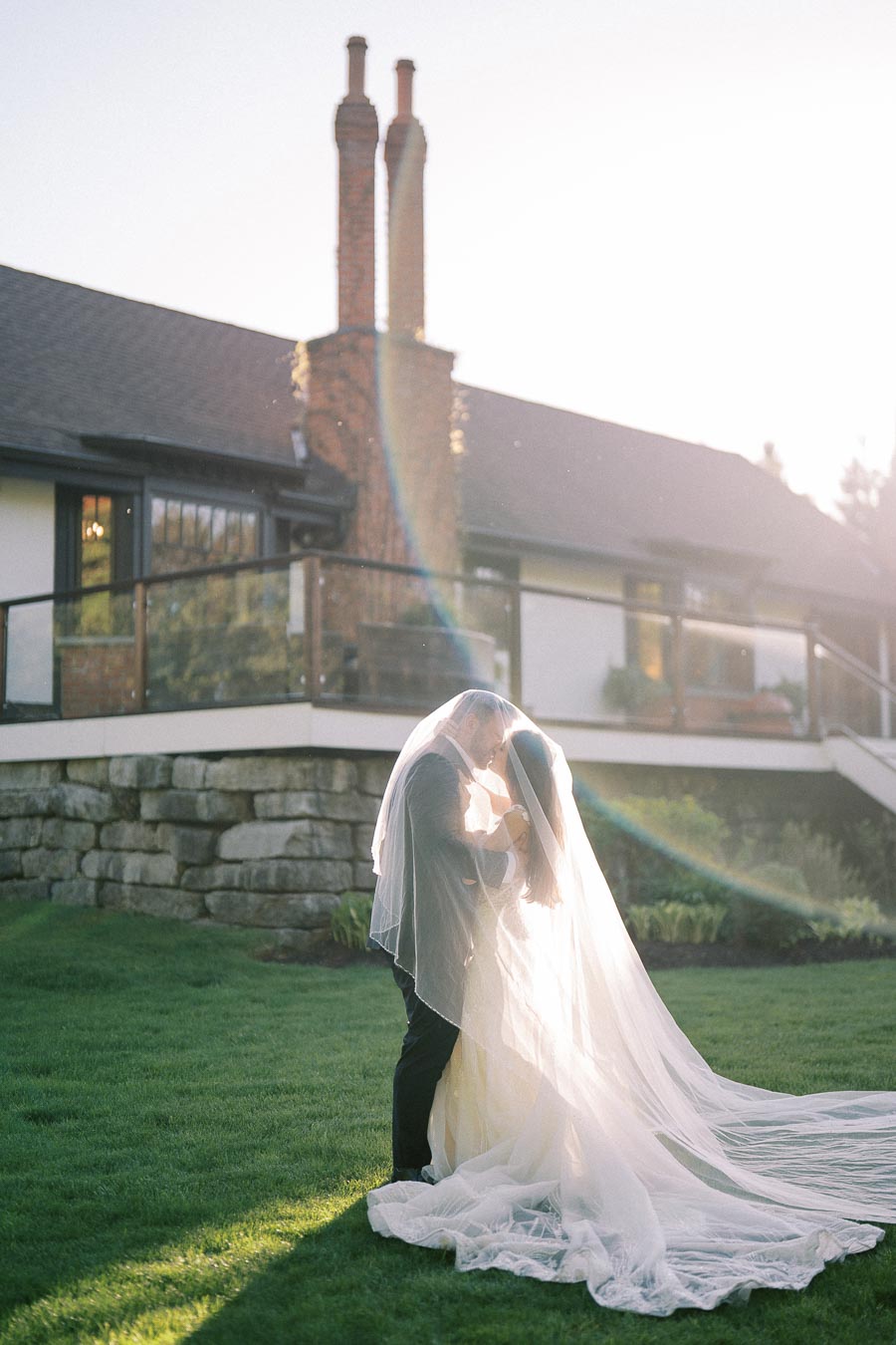 A bride and groom share an intimate moment under a sheer veil, standing in a sunlit garden in front of a rustic house.