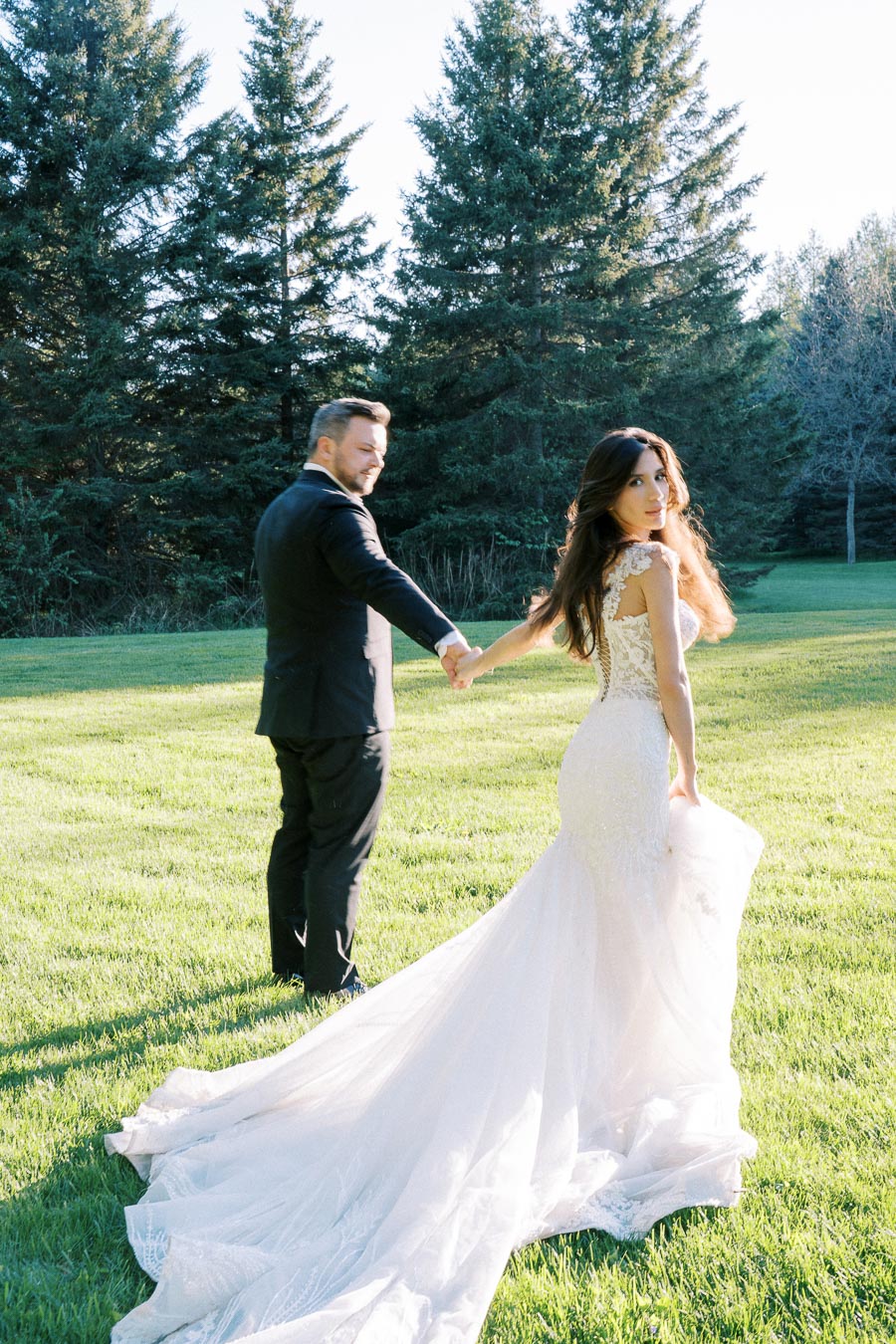 A bride in a stunning lace wedding dress holding hands with the groom in a black suit, walking on lush green grass with tall evergreen trees in the background.