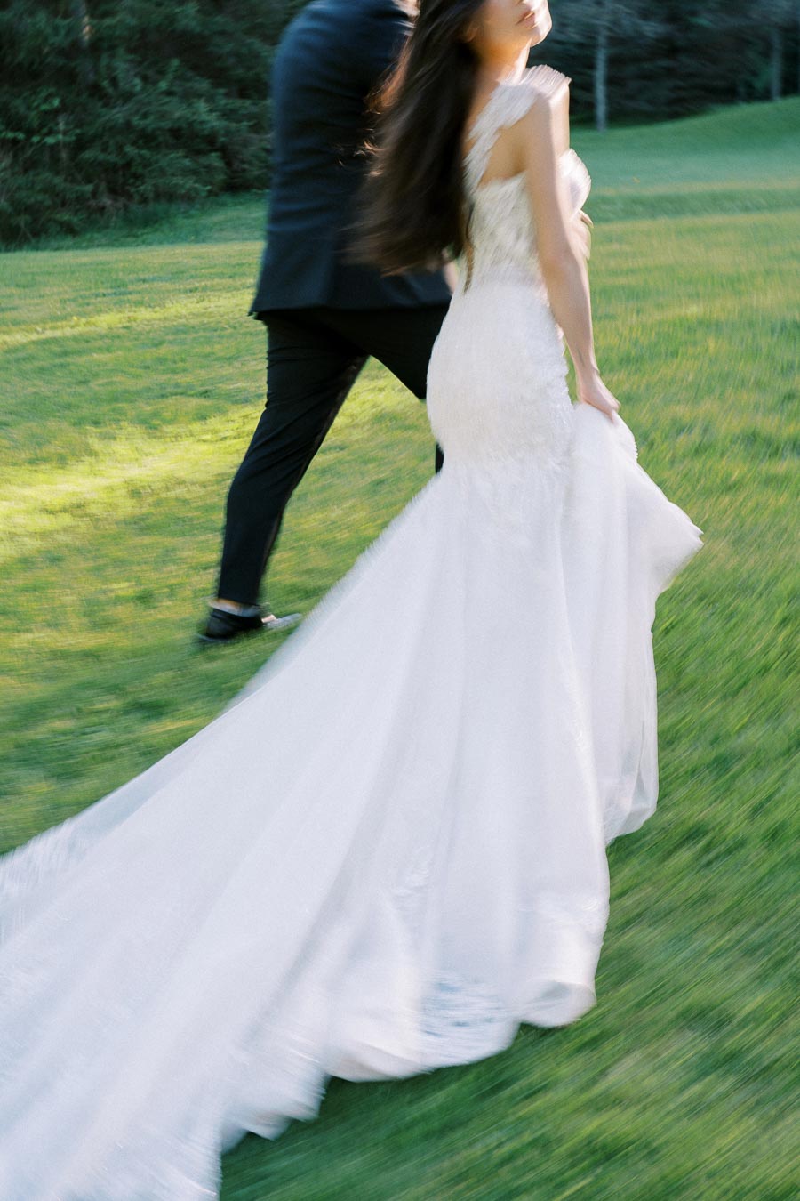 Blurred image of a bride in an elegant white gown walking with a groom on a grassy lawn.