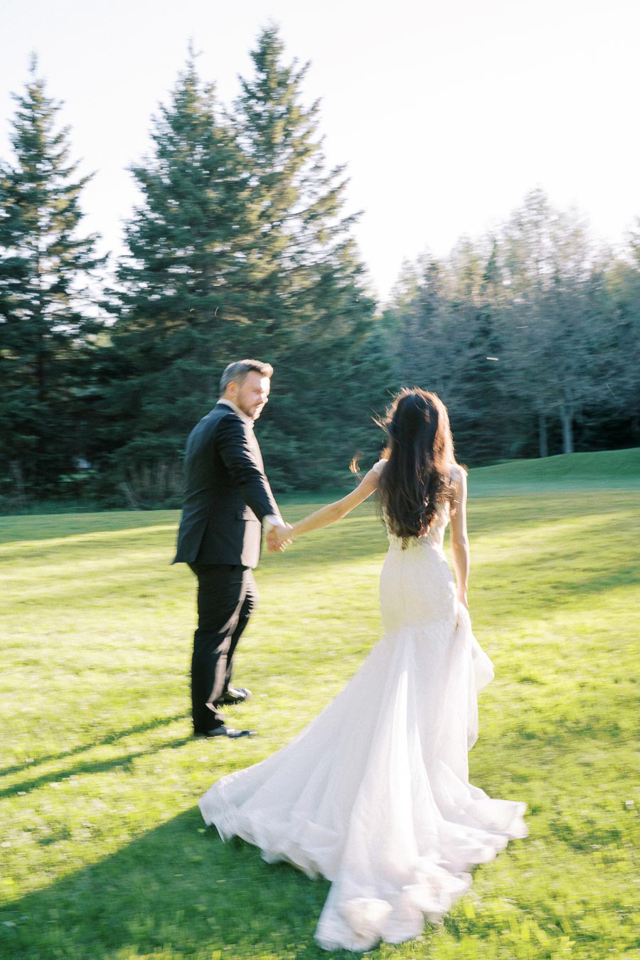 A bride and groom holding hands and walking on a sunlit grassy field surrounded by tall trees, captured during a romantic outdoor wedding.
