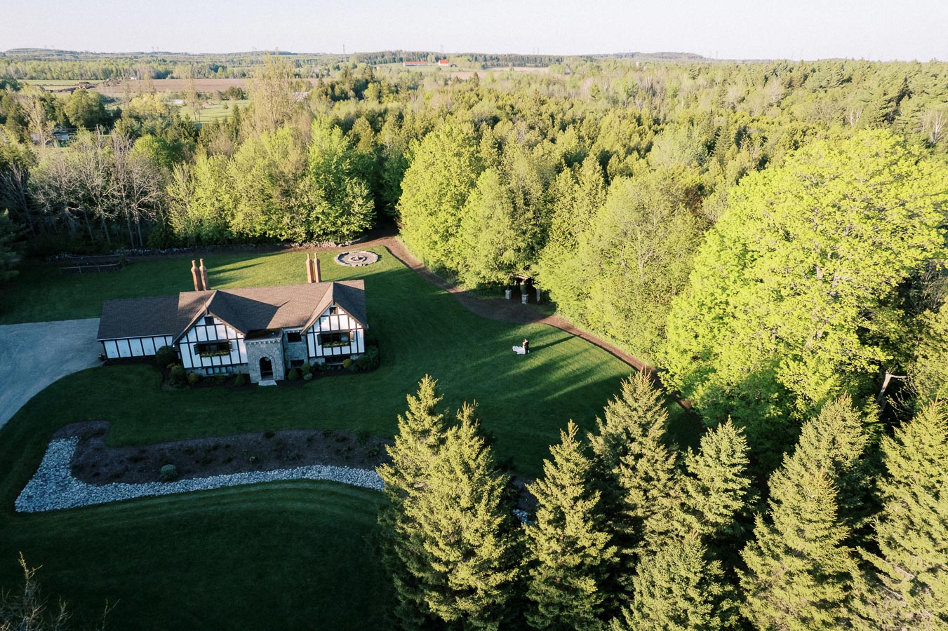 Aerial view of a charming countryside estate surrounded by lush green trees, featuring a traditional house with a spacious lawn and picturesque landscape.