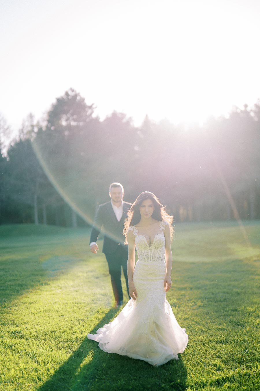 Romantic wedding photo with bride in lace gown and groom in suit, walking on a sunlit grassy field backed by a forest, capturing ethereal sunlight and natural beauty.