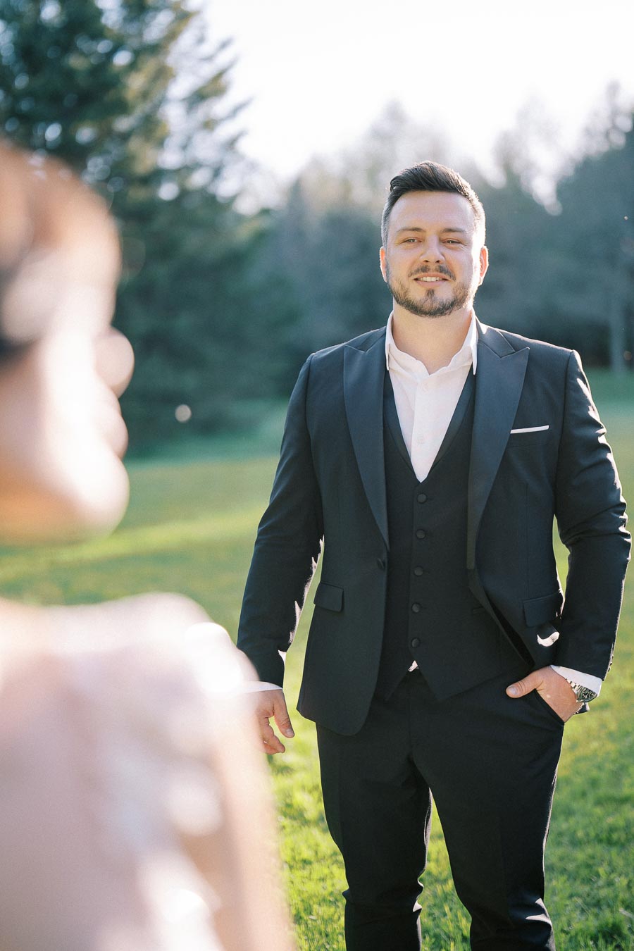 A man in a formal suit posing outdoors in a sunny garden setting, with trees and greenery in the background, exuding a confident and stylish appearance.