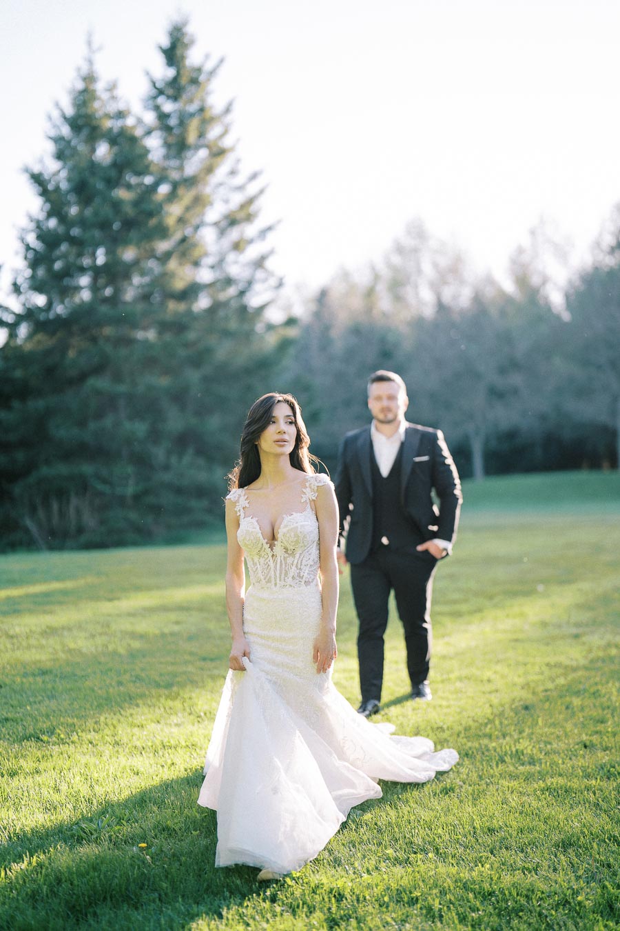 Bride in elegant lace wedding dress walks on a sunlit lawn, followed by groom in a dark suit with a forest backdrop.
