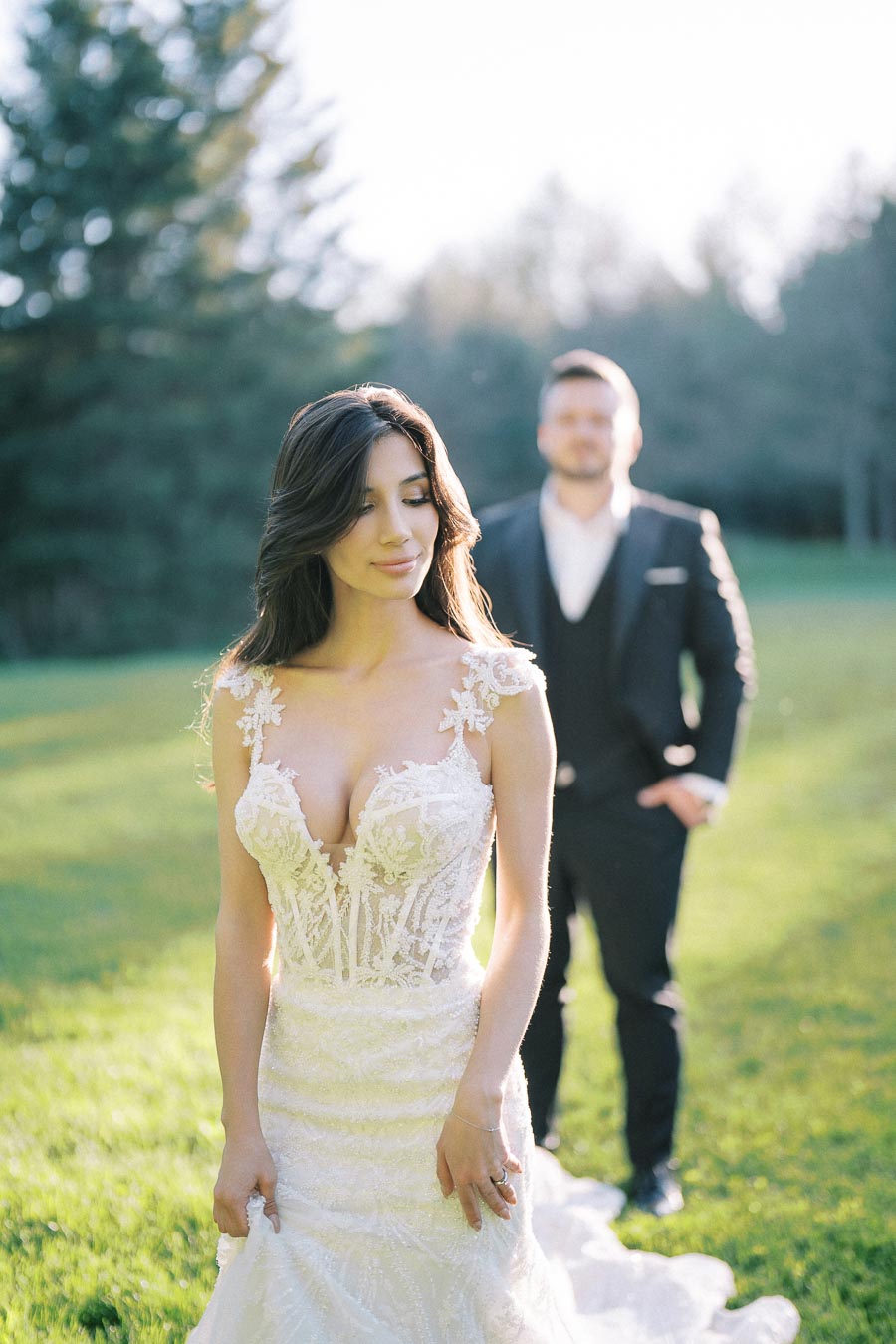 A bride in a lace wedding gown stands in a sunlit garden, with a groom in a dark suit standing in the background, surrounded by lush greenery.