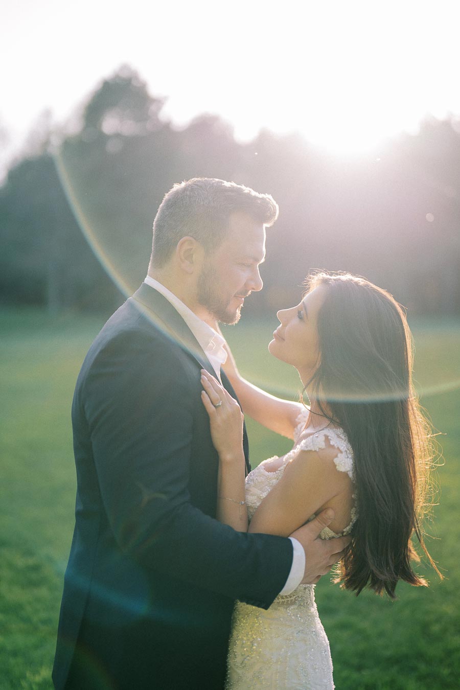 Romantic couple in formal attire embracing outdoors at sunset, with sun flare enhancing the background, capturing a tender moment of love and connection.