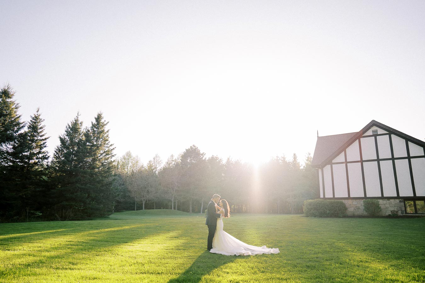 A bride and groom embrace on a sunny lawn with a picturesque house and tall evergreen trees in the background, bathed in warm sunlight.